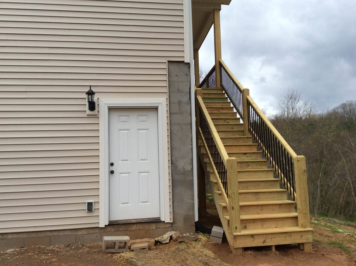 Wooden exterior staircase with black metal railing beside white door featuring black hardware, stone siding, and wall-mounted lantern under cloudy sky.
