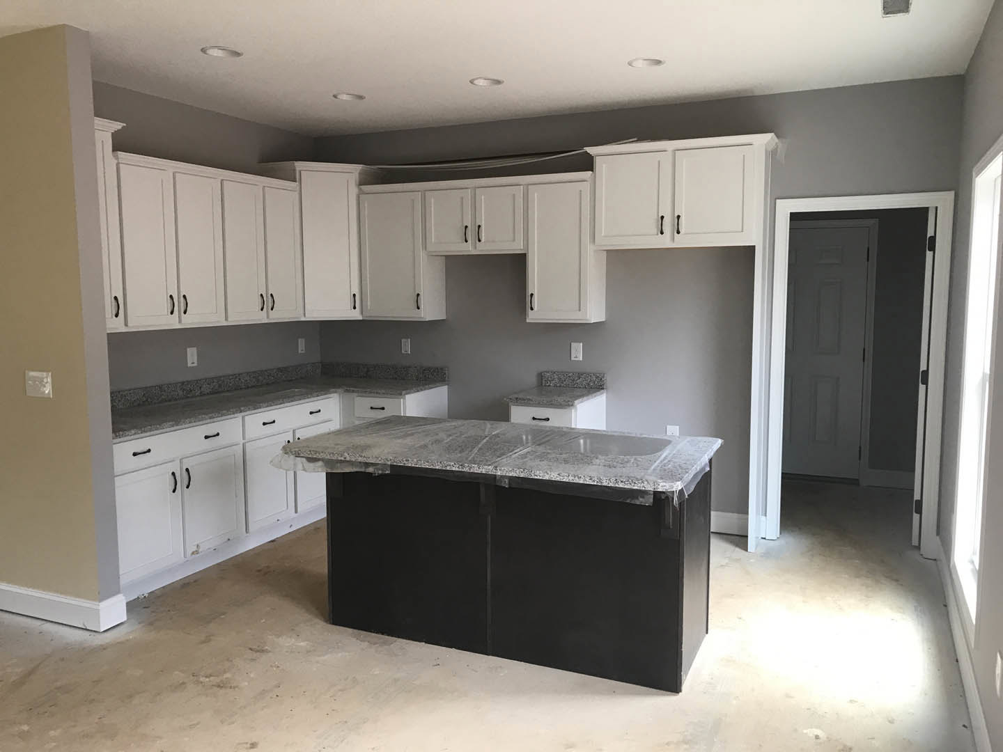 White kitchen cabinets and black island with marble countertop partially wrapped in plastic, stainless steel sink, white tile backsplash, black door with white frame, recessed