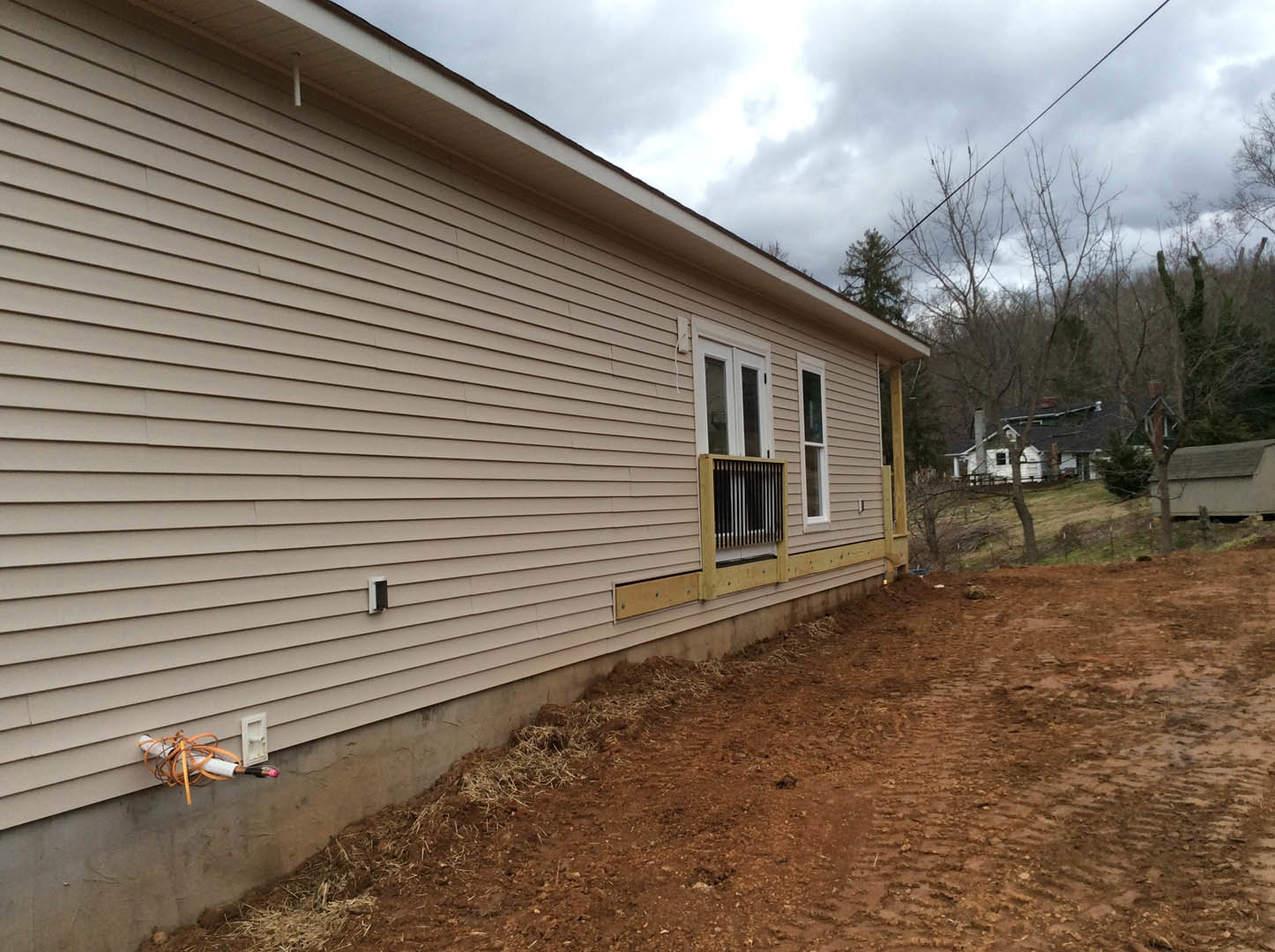 Two-story home with light siding, barred window, exposed pipe, and dirt patch beside the foundation; trees and cloudy sky in background.