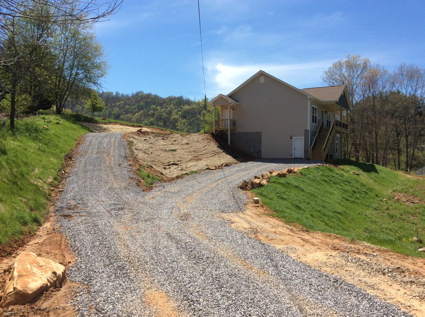 Gravel driveway curving toward a two-story house with balcony, surrounded by grassy hill, large rock, and cluster of trees under partly cloudy sky