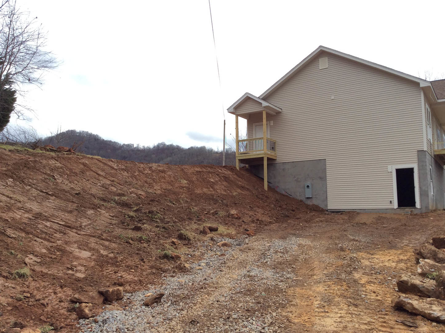 Two-story cottage with white siding, black front door, covered porch, bare tree, dirt hill in foreground, power line overhead, and rocky landscaping.