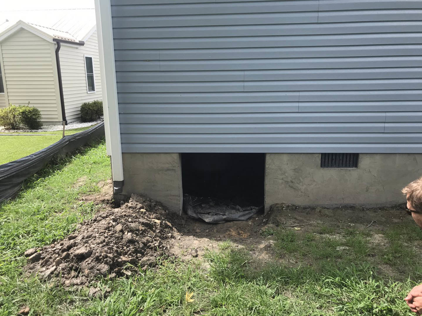 White house exterior with attached garage, black gutter, and green grass lawn partially covered by a black tarp; basement foundation visible below ground level.