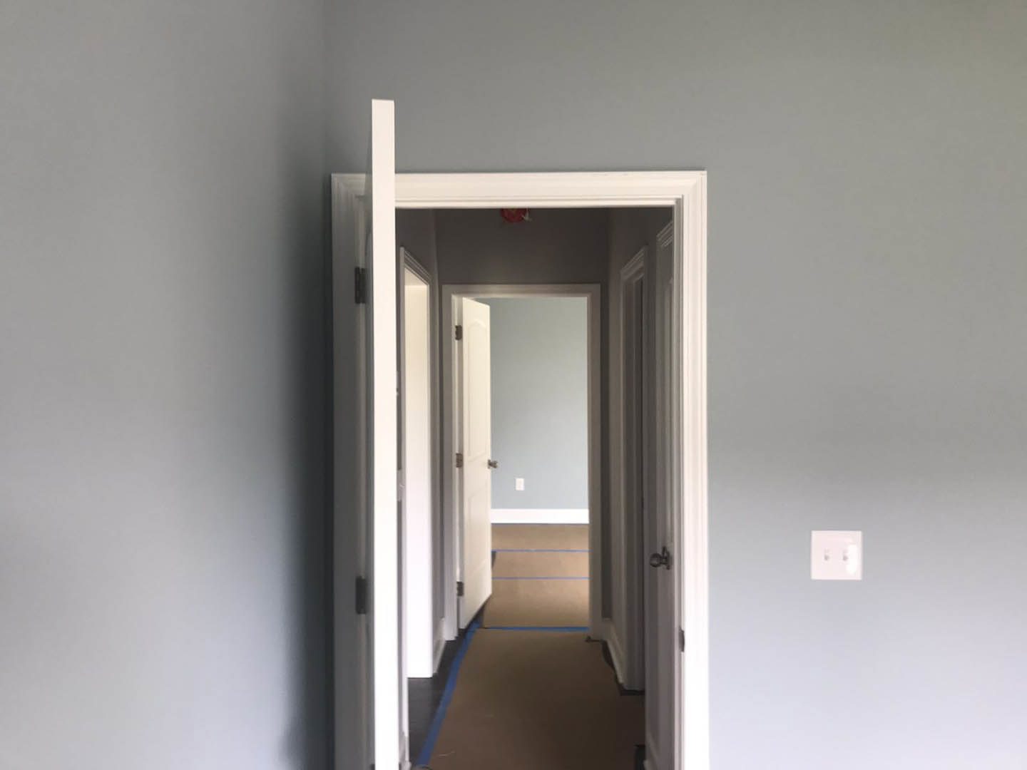 Hallway with white paneled doors, blue carpet flooring, white walls with black trim, silver door knobs, blue painter’s tape, and a white double light switch.