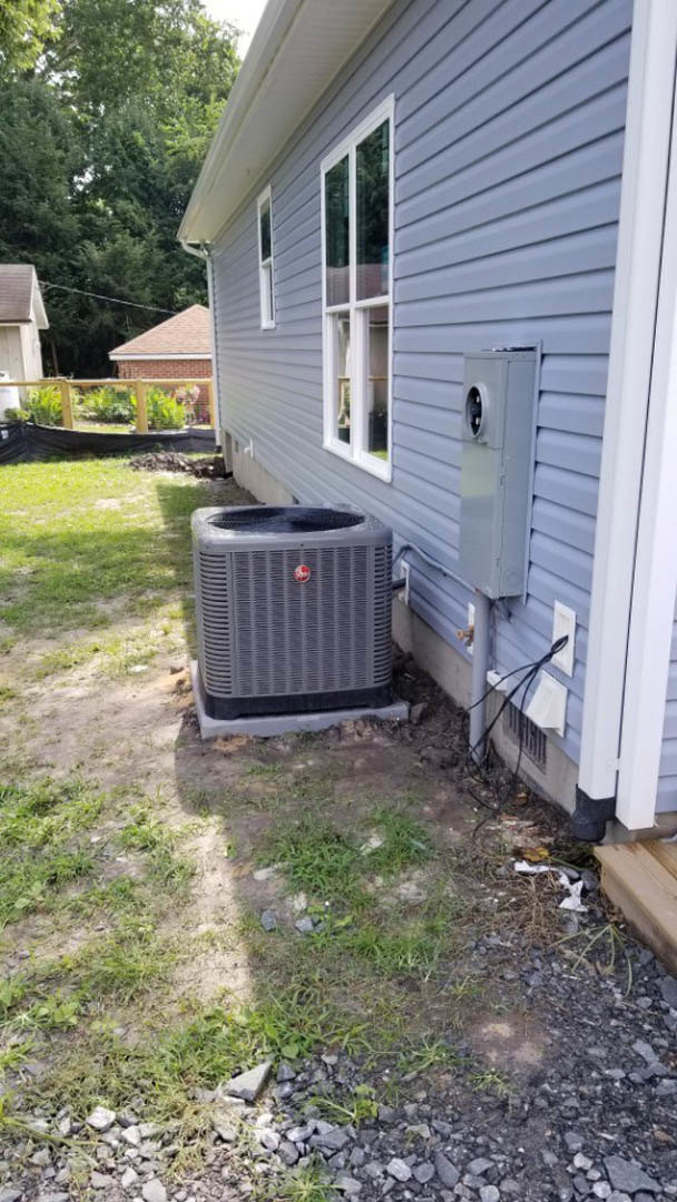 Grey heat pump unit on concrete pad beside brick house with white door and window, surrounded by grass and plants in backyard.
