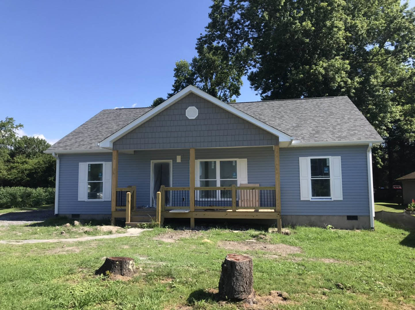 White house with covered porch, wooden railing, white shutters, fenced yard, tree stump on grassy lawn, small cottage visible in background.