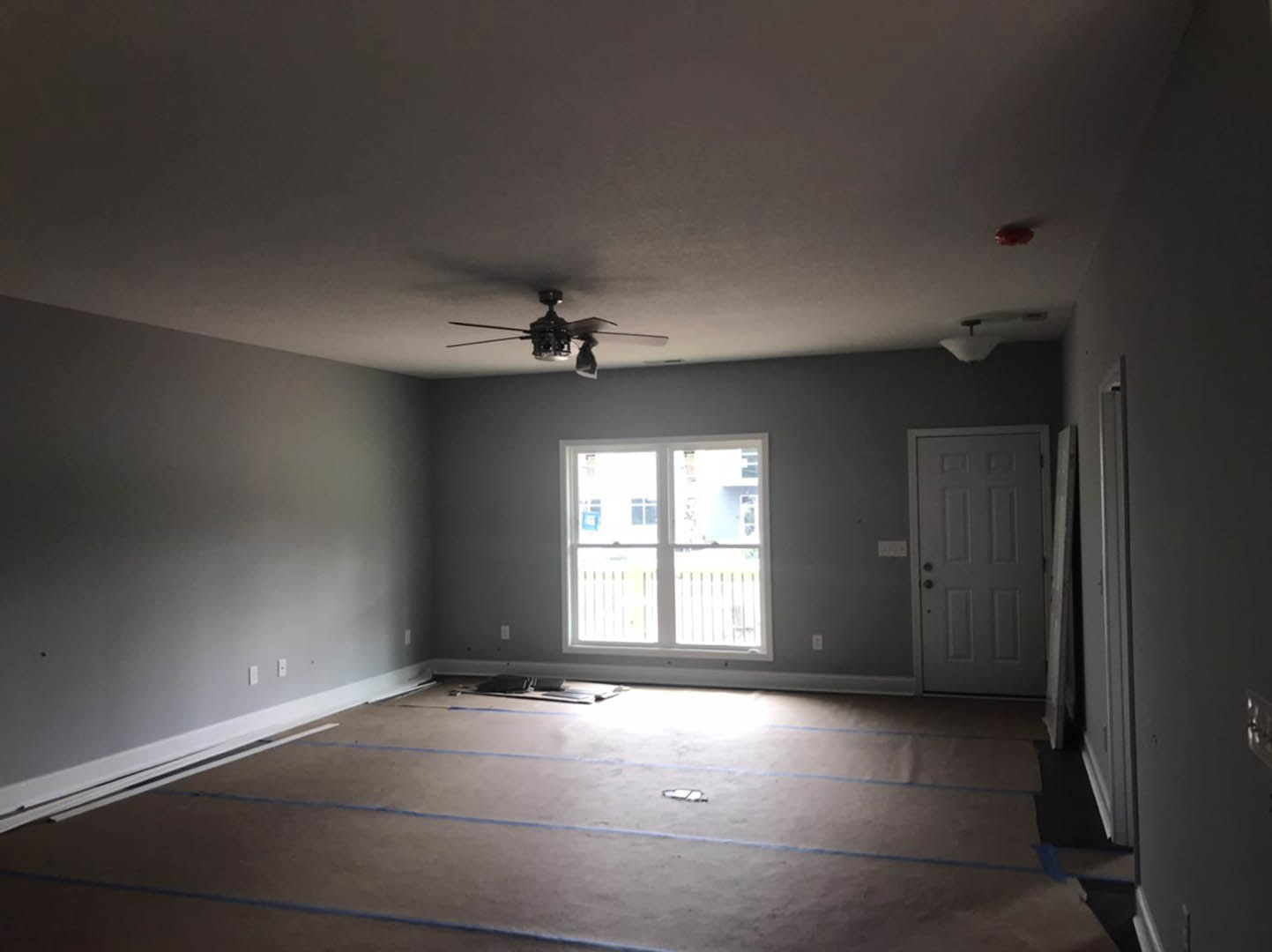 Carpeted room with white-framed window, ceiling fan with light fixture, white walls, and blue tape marking sections on the floor
