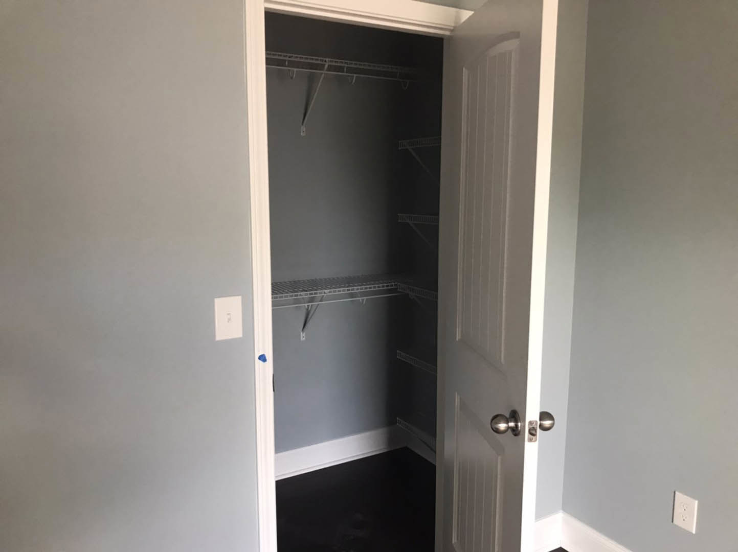 Closet with built-in white shelves, white walls, black flooring with white trim, white electrical outlet, close-up of brushed metal door knob on white door
