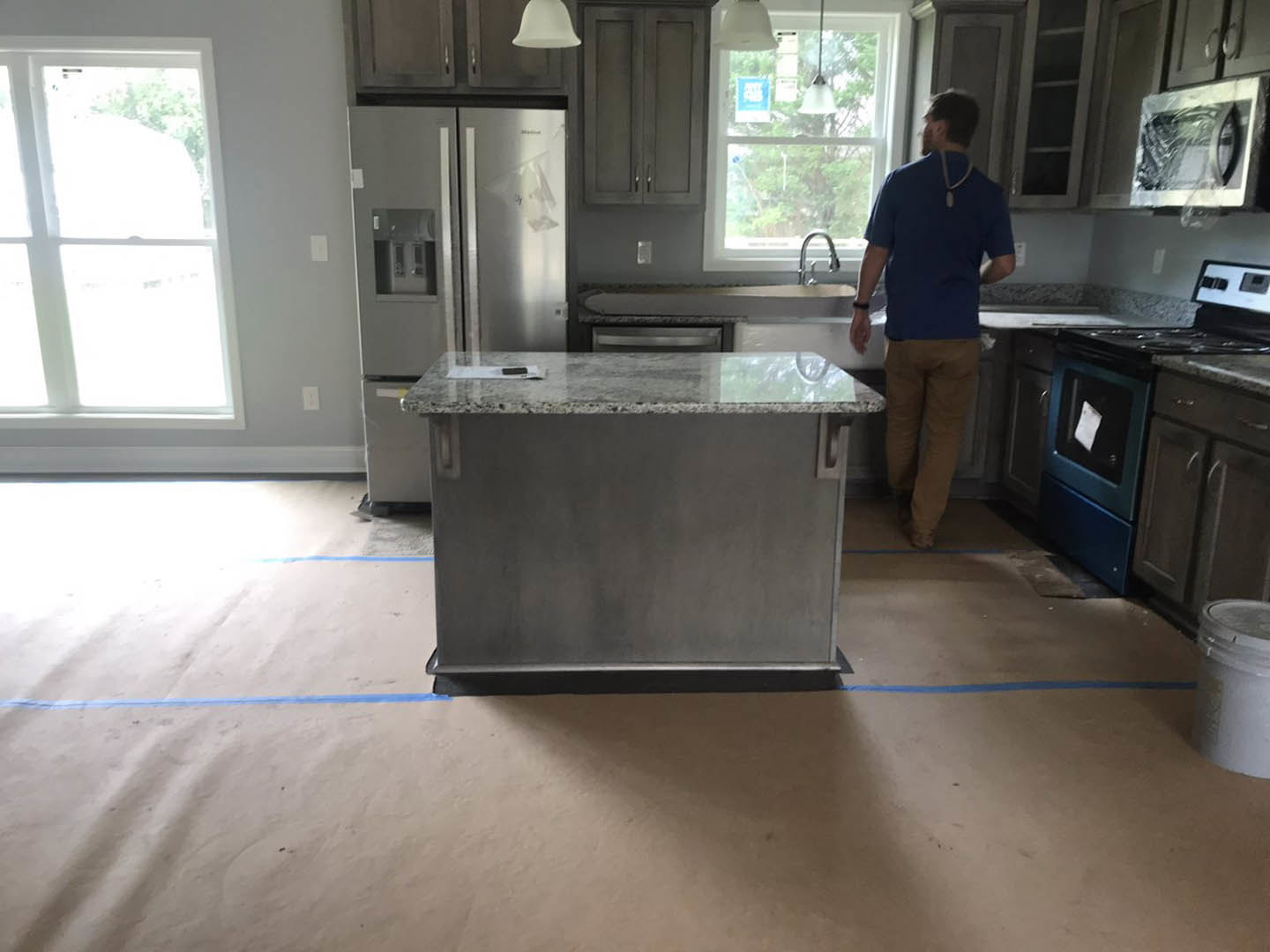 Man in blue shirt standing in kitchen with marble countertop, white cabinetry, stainless steel appliances, window with white frame, white bucket with lid, and piece of paper on