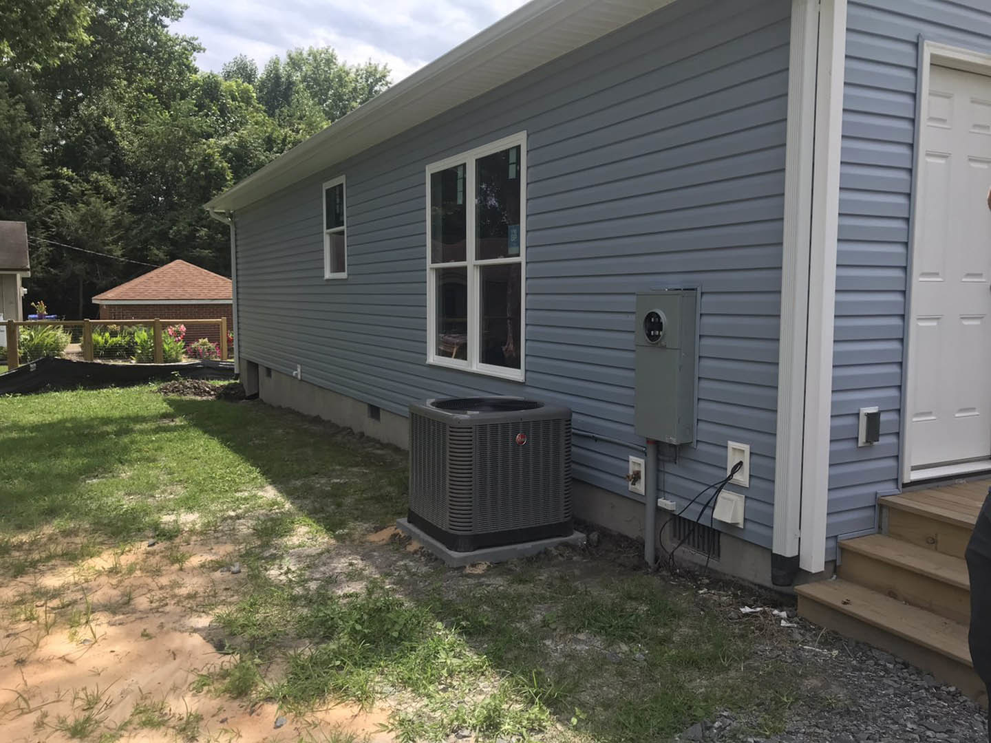 Gray heat pump mounted on concrete base beside brick house with white-framed window, white door, fenced backyard, and green grass