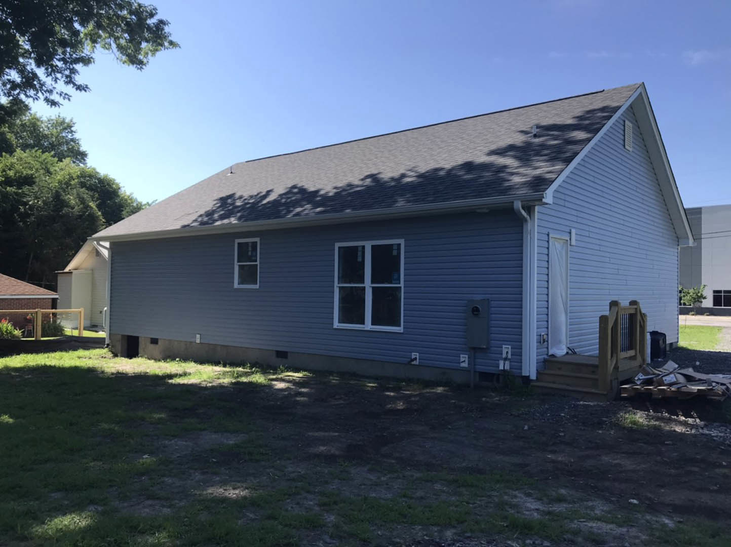 Two-story house with blue metal roof, white siding, covered front porch, white-framed windows, dirt patch and grass in front yard, tree shadows cast on roof and wall.