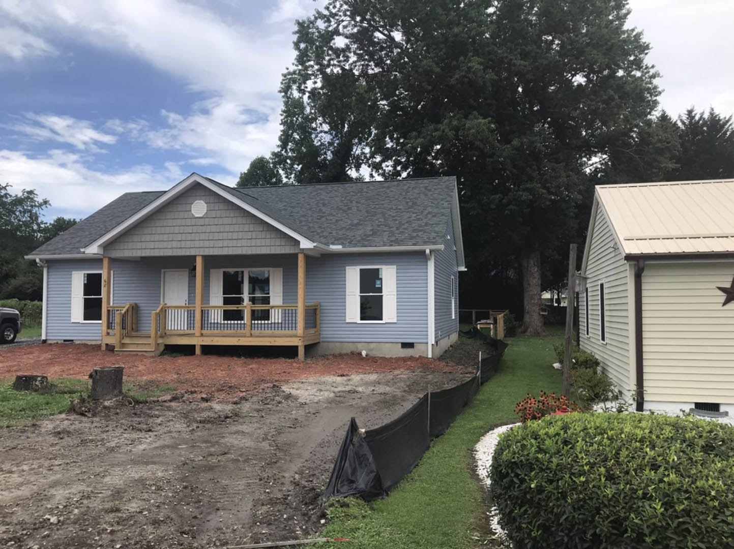 Two-story cottage with white-framed windows, wooden porch and deck, fenced yard featuring green grass, bushes, and a tree stump under a partly cloudy sky