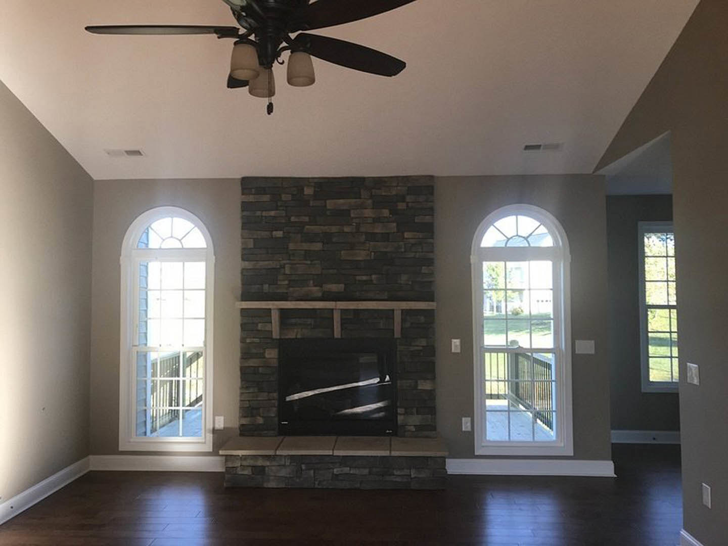 Living room with brick fireplace, large windows, dark carpet flooring, ceiling fan, and white wall molding