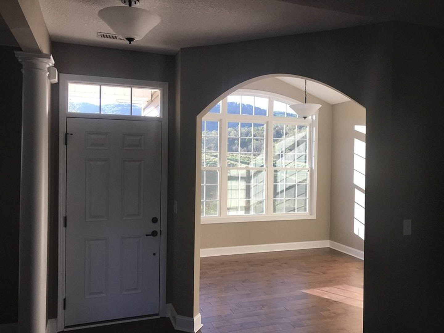 White door with black handle, large window framing mountain view, wooden floor, white lamp shade, person standing near metal pole