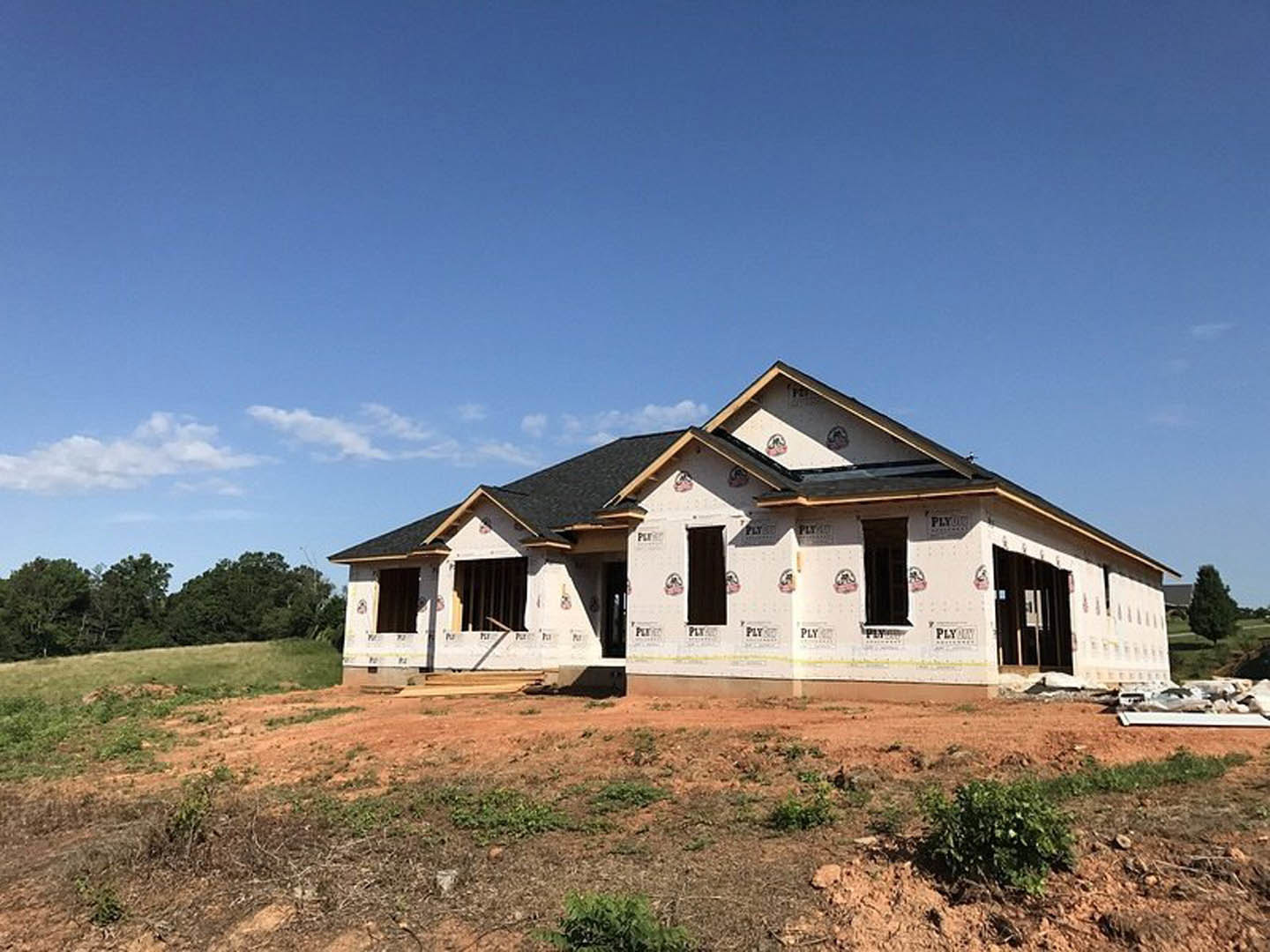 Two-story house under construction with white exterior walls, surrounded by a dirt field and scattered trees under a cloudy sky