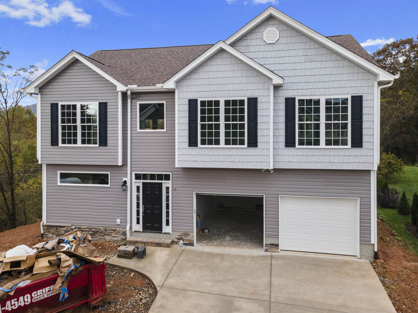 Two-story house with white siding and black-trimmed windows, attached garage with white door, black front door with white trim, red dumpster filled with cardboard on driveway