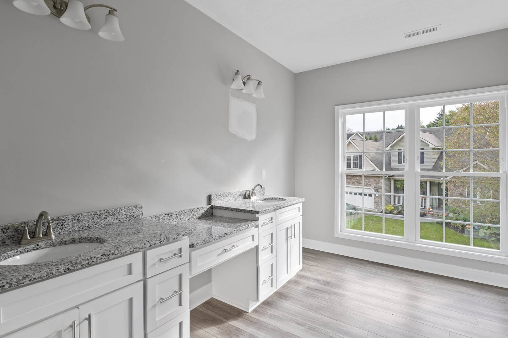Bathroom with marble countertops, undermount sink, three-light fixture above mirror, chrome faucet, window with view of neighboring house, white cabinetry, and neutral wall