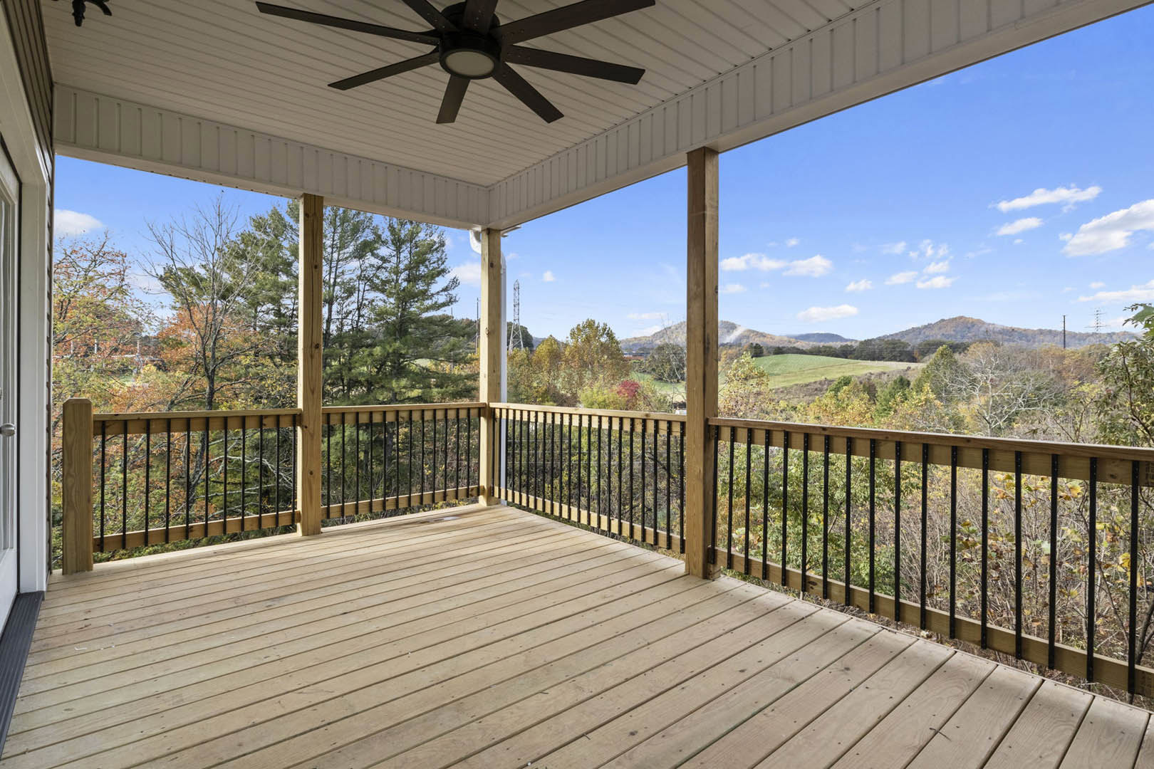 Wooden deck with black metal railing, ceiling fan with light fixture overhead, surrounded by leafy trees