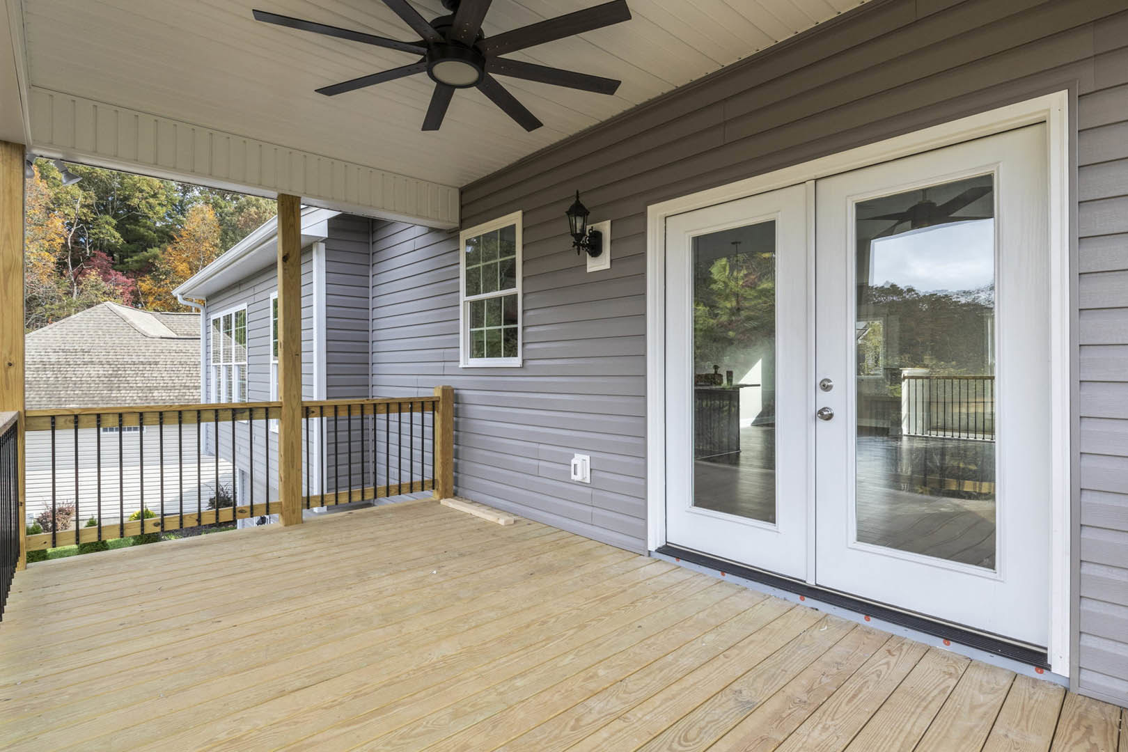 Wooden deck with white double doors featuring glass panes, ceiling fan with light, white-framed window, and close-up of railing
