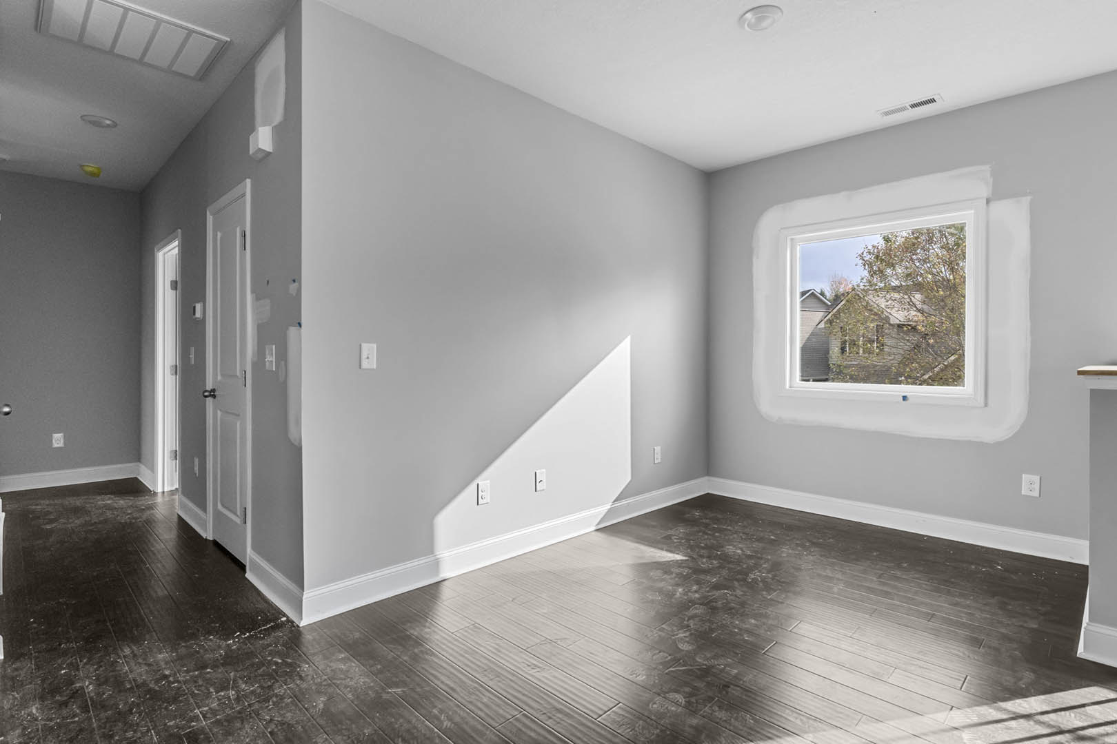 Wood flooring and white walls in a bright room with a large window, tree and house visible outside, white ceiling with rectangular pattern, and white door with silver knob