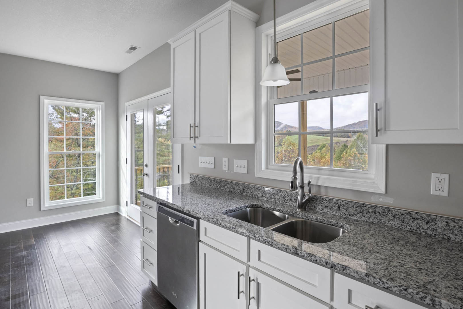White kitchen cabinets and granite countertops, stainless steel sink with chrome faucet, grey wood flooring, large window overlooking trees, modern pendant light fixture, white