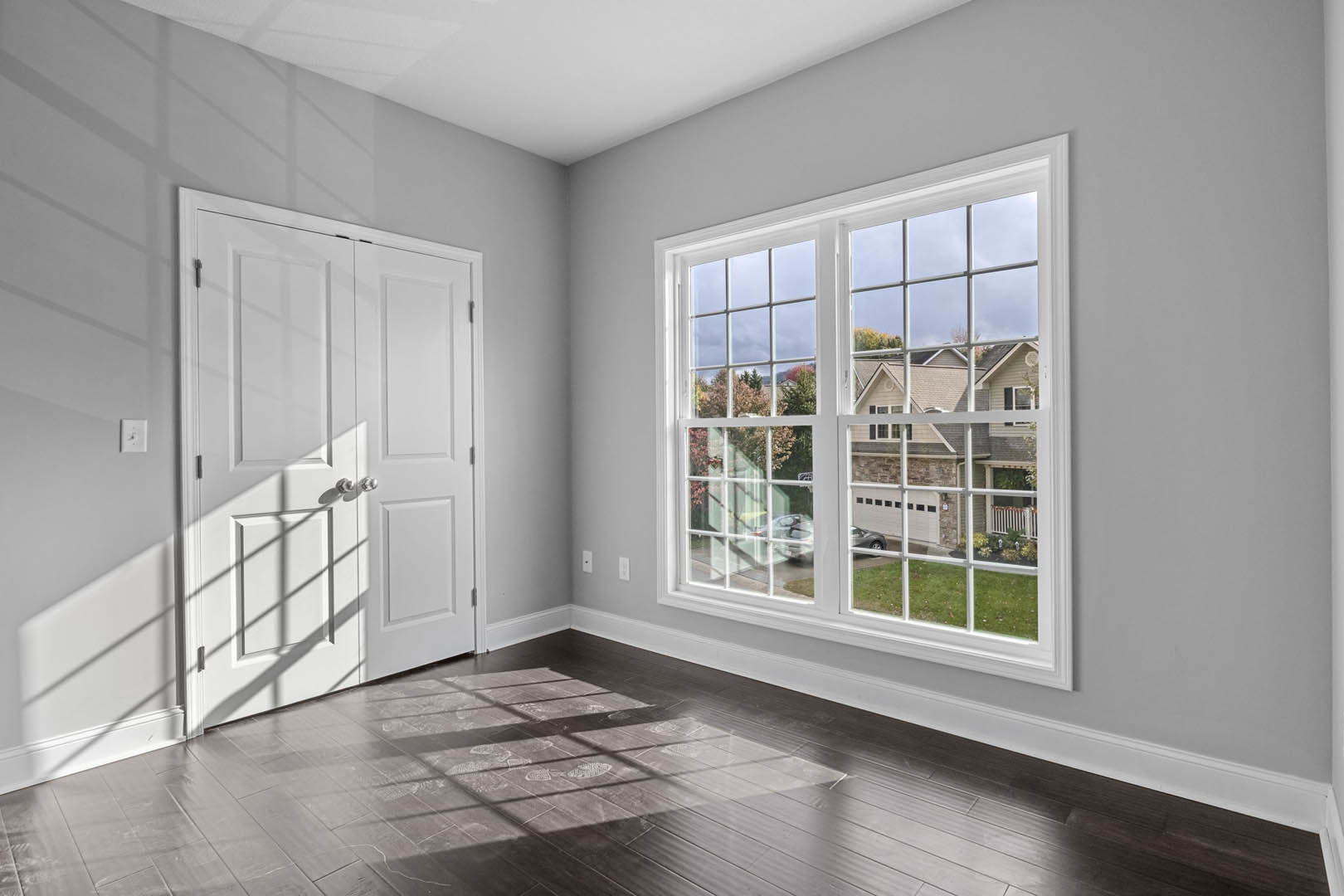 Sunlit room with white plaster walls, wood flooring, large window casting shadows, white door with silver knobs, view of neighboring house and parked car through window