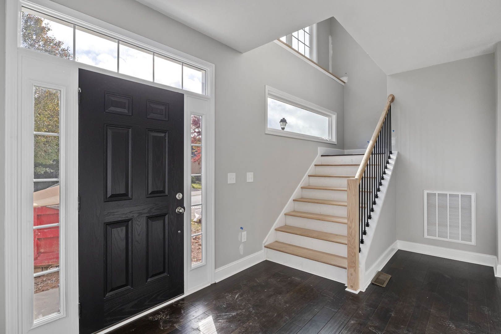 Wood staircase with white risers ascending toward a matte black door, dark brown tile flooring, white wall vent, window with lamp on sill, plaster walls.
