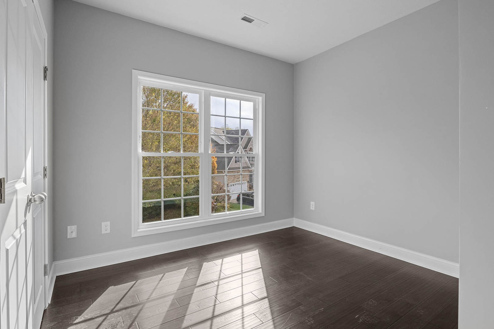 Sunlit room featuring a large multi-pane window overlooking trees and neighboring houses, hardwood flooring, white plaster walls, and a ceiling vent.