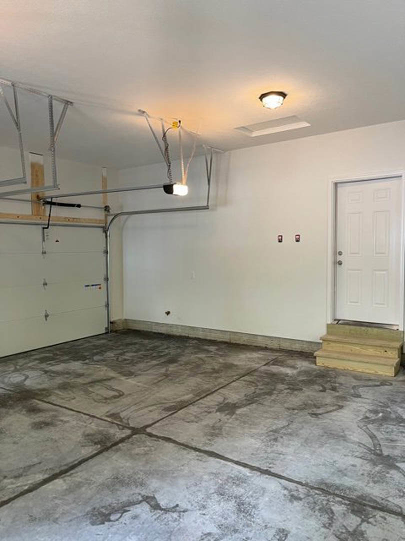 Garage interior with white entry door featuring silver knobs, concrete floor, wooden step, plaster walls, and ceiling-mounted light fixture