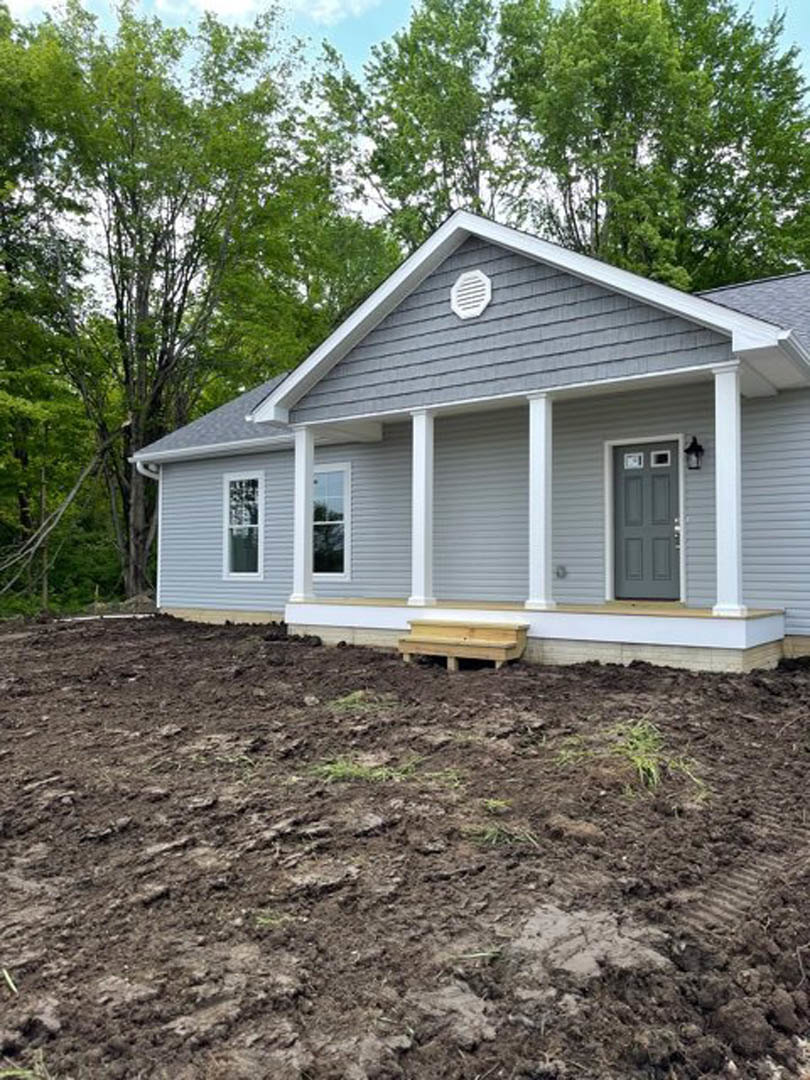 Two-story home with gray siding, white trim, covered porch, vented roof, and muddy yard with scattered benches.