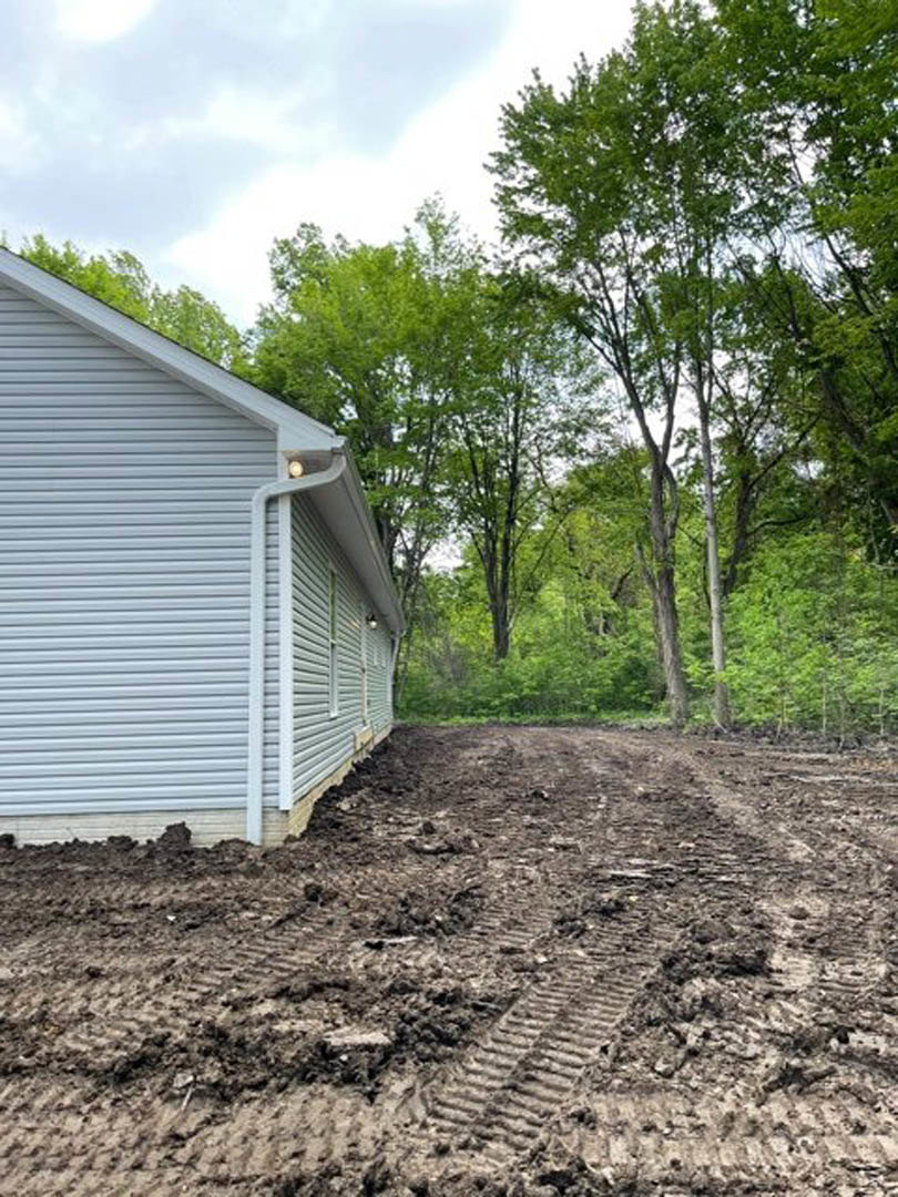 Side view of a custom home with muddy ground and tire tracks, surrounded by trees and cloudy sky