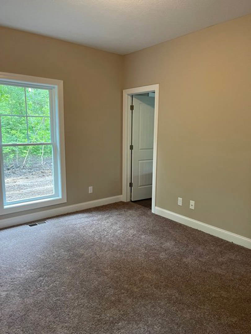 Neutral-toned room with laminate flooring, open white door featuring a metal bar, large window overlooking dense forest, light-colored carpet, plaster walls