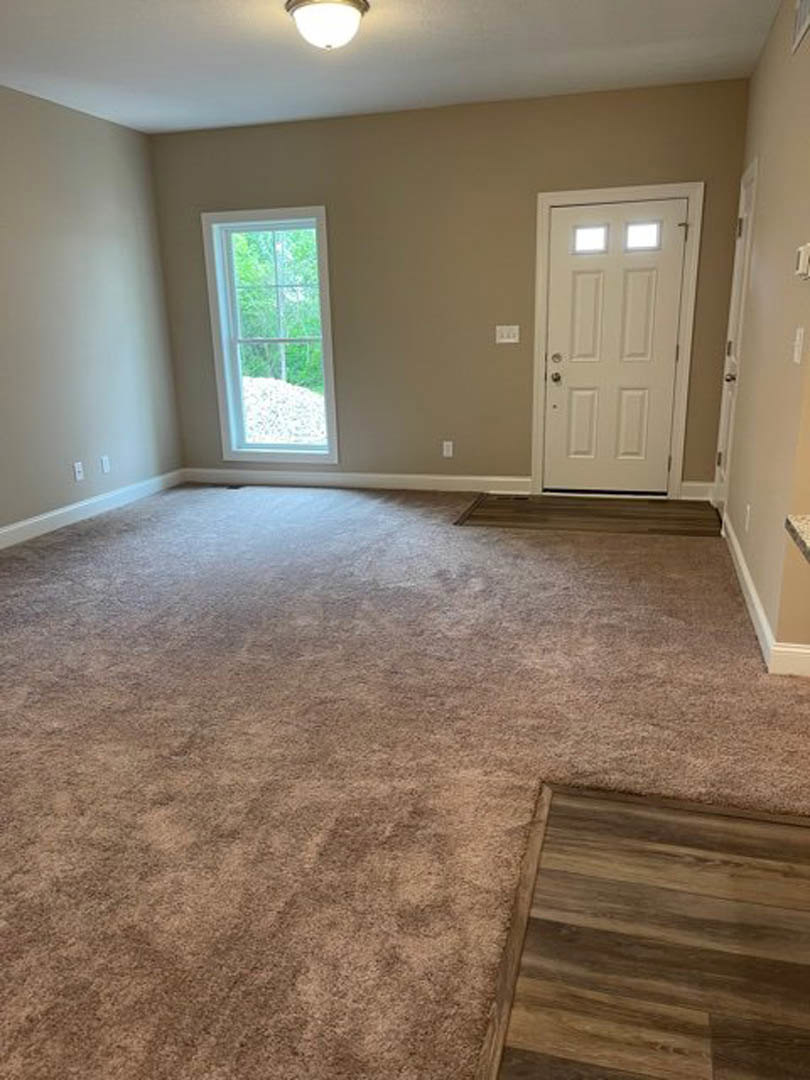 White paneled door with glass inserts, beige carpeted floor, wood flooring beneath area rug, large window overlooking trees, white plaster walls, ceiling-mounted light fixture