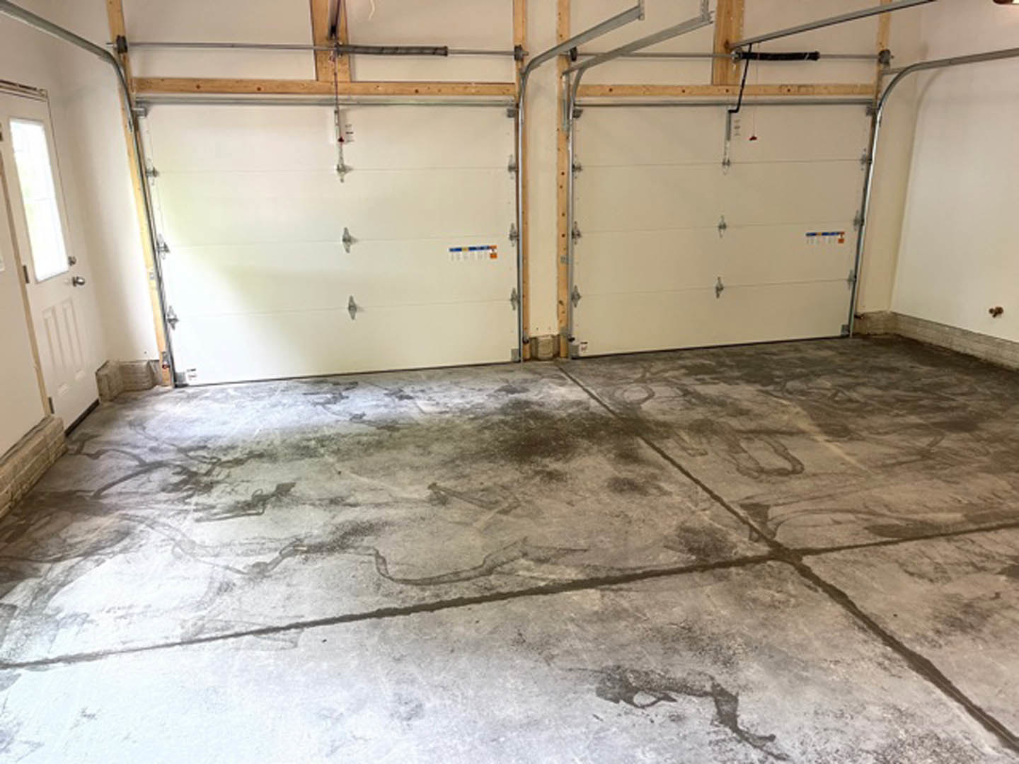 Garage interior with smooth concrete floor, white paneled door featuring metal handles and bars, plaster walls, and a close-up window; couch visible in background.