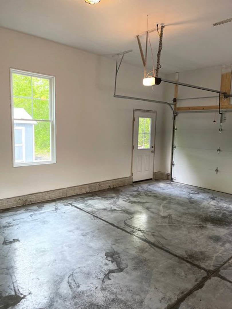 Garage interior with smooth concrete floor, white plaster walls, white door with window, large window framed in white showing neighboring house, exposed wire near ceiling