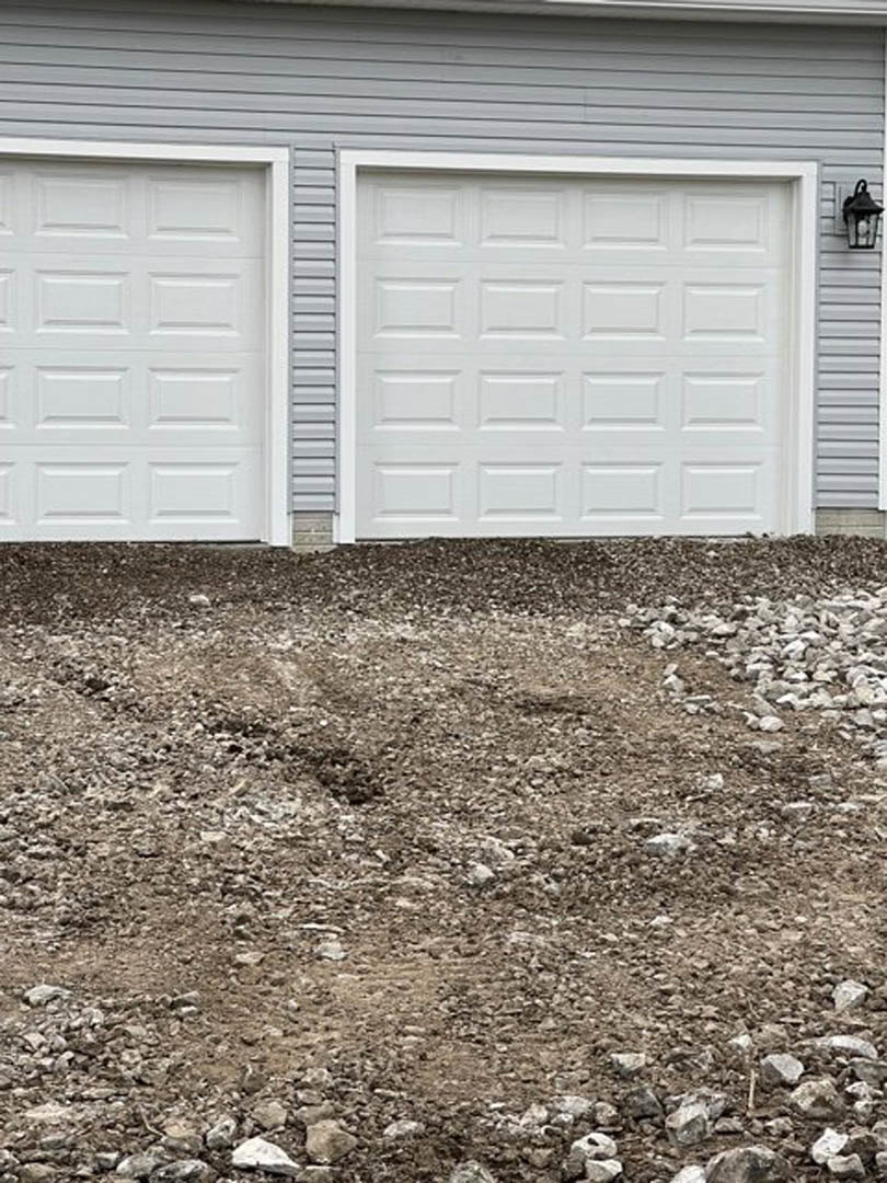 White paneled garage door with black trim, exterior wall-mounted light fixture on the side, dirt and rocks covering unfinished yard.