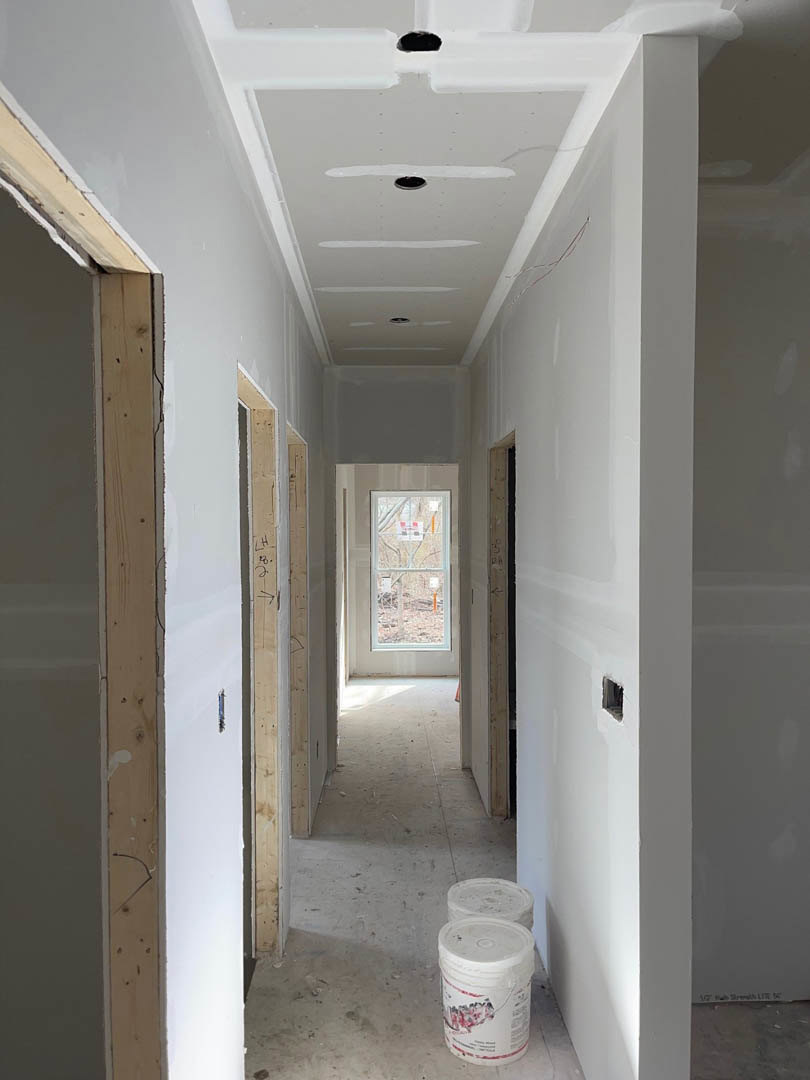 Hallway with smooth white plaster walls and ceiling, recessed lighting holes, white-framed window, exposed wood beam detail, paint bucket on floor, and white trim along wall edges