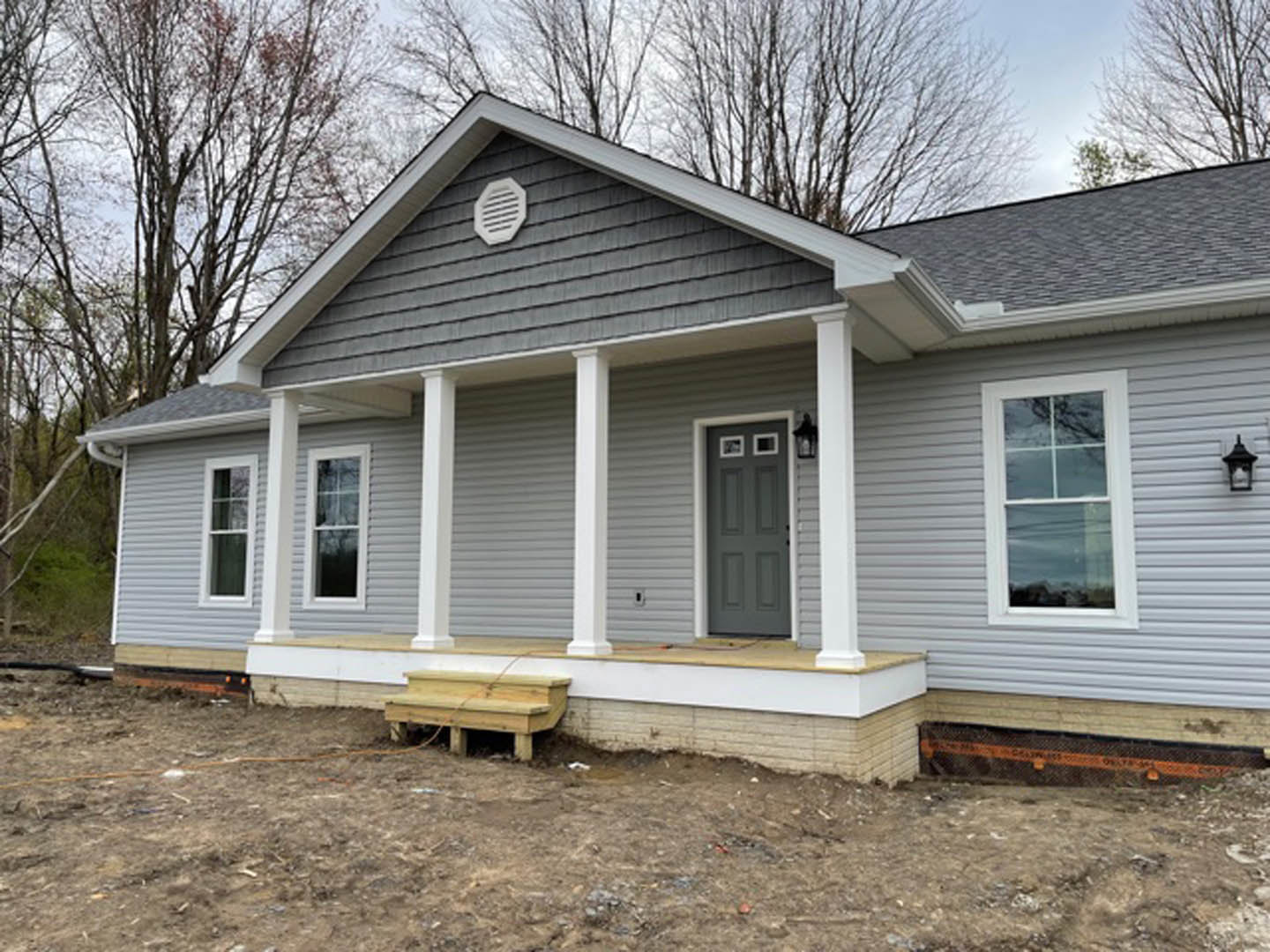Grey house under construction with white pillars, grey door with white trim, white-framed window, roof vent, exposed wooden structure, surrounded by dirt field and trees.