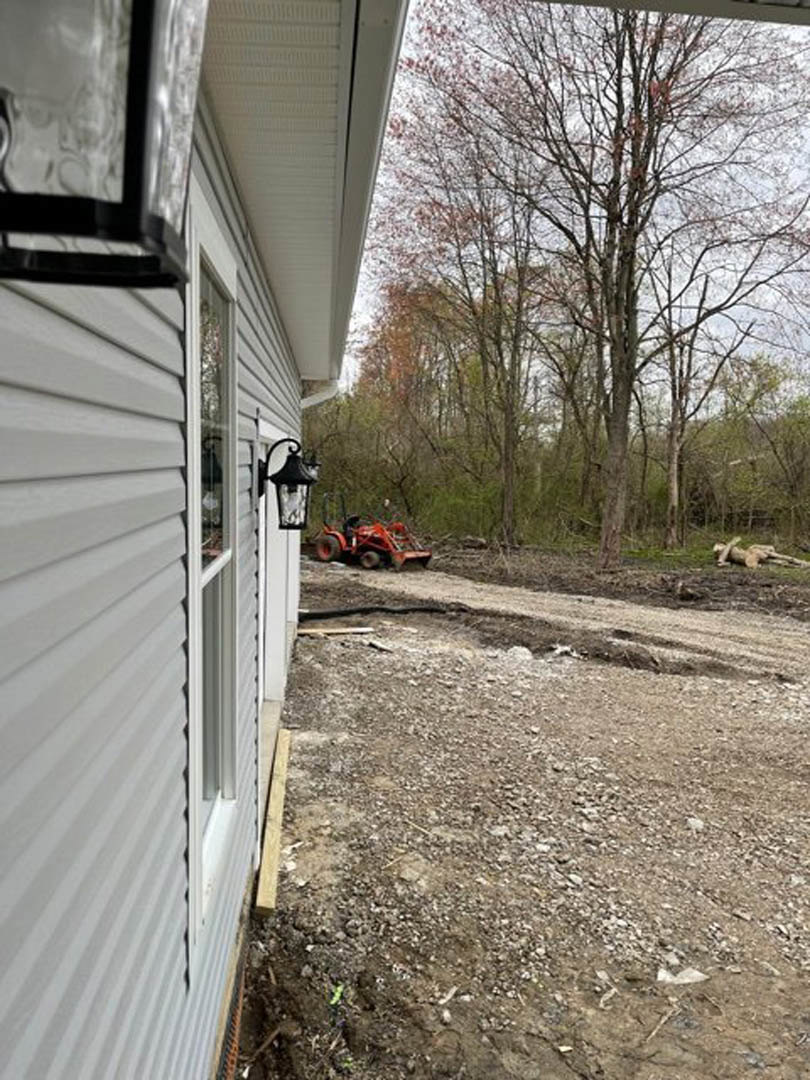 Red tractor parked on dirt driveway beside white house with horizontal siding, surrounded by trees and greenery under a blue sky