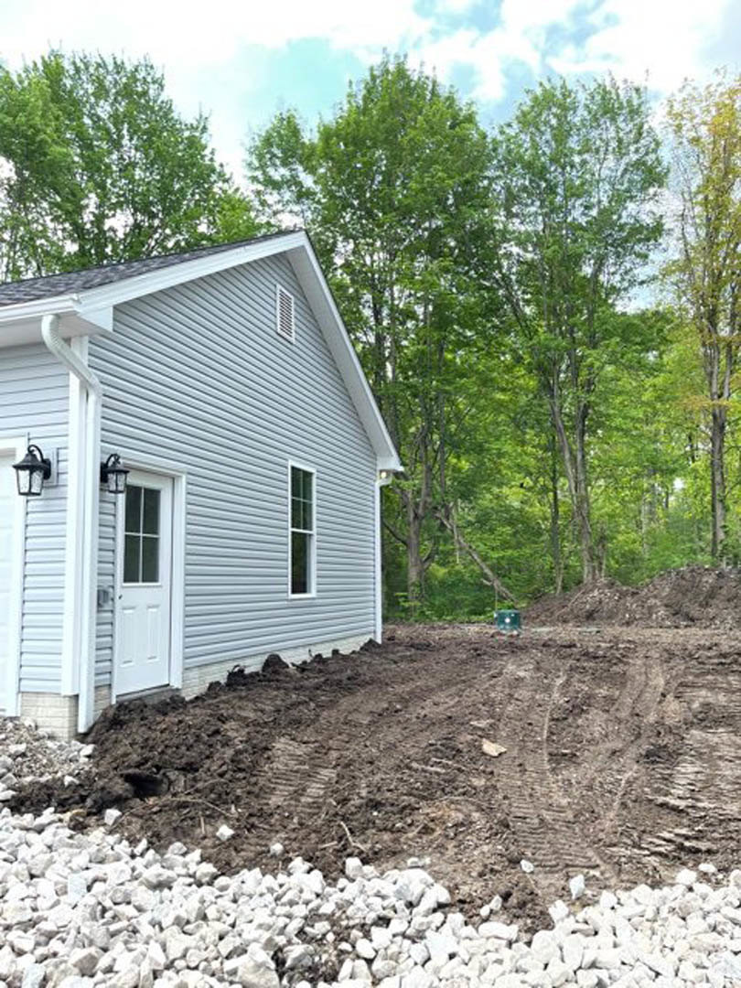 Two-story home with light siding, white front door, black light fixture, dirt driveway, and mature trees in the background