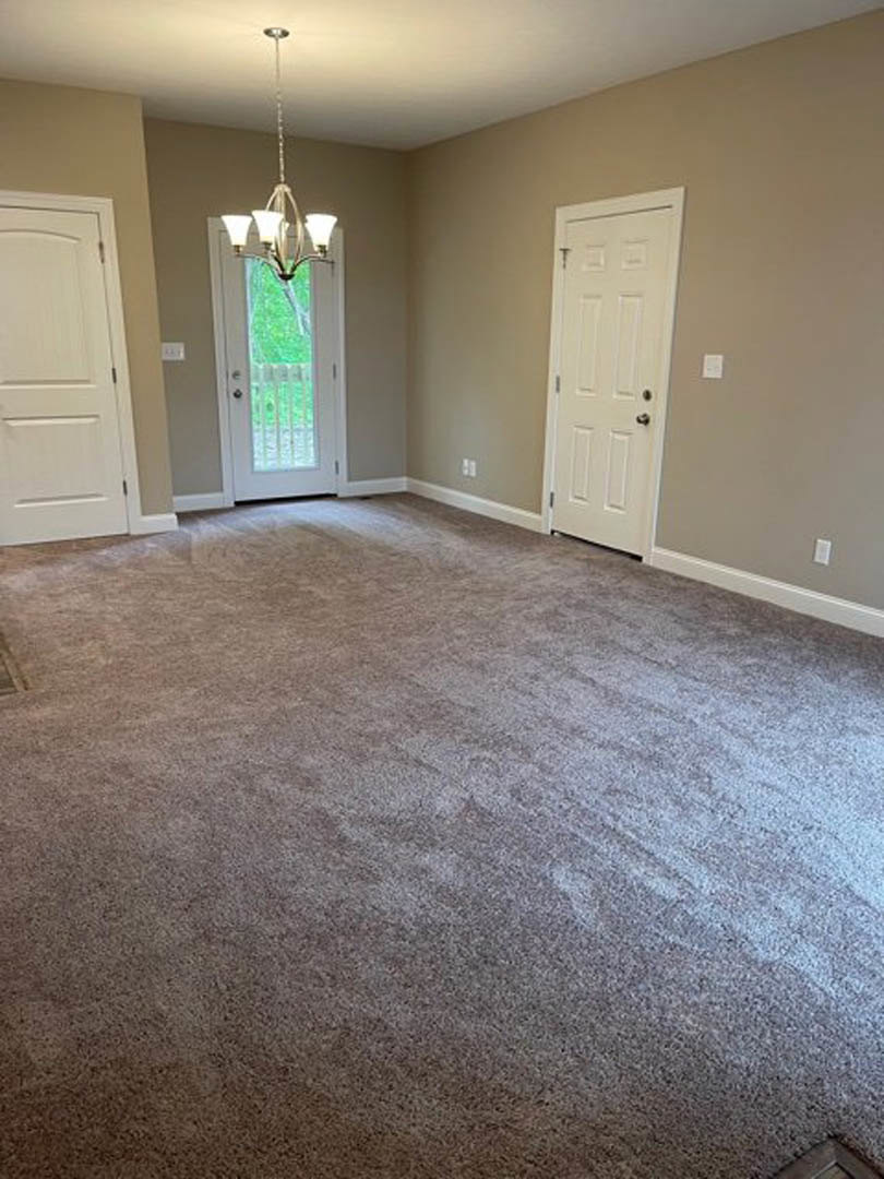Carpeted room with white paneled door, silver handle, two windows, and modern chandelier hanging from white plaster ceiling