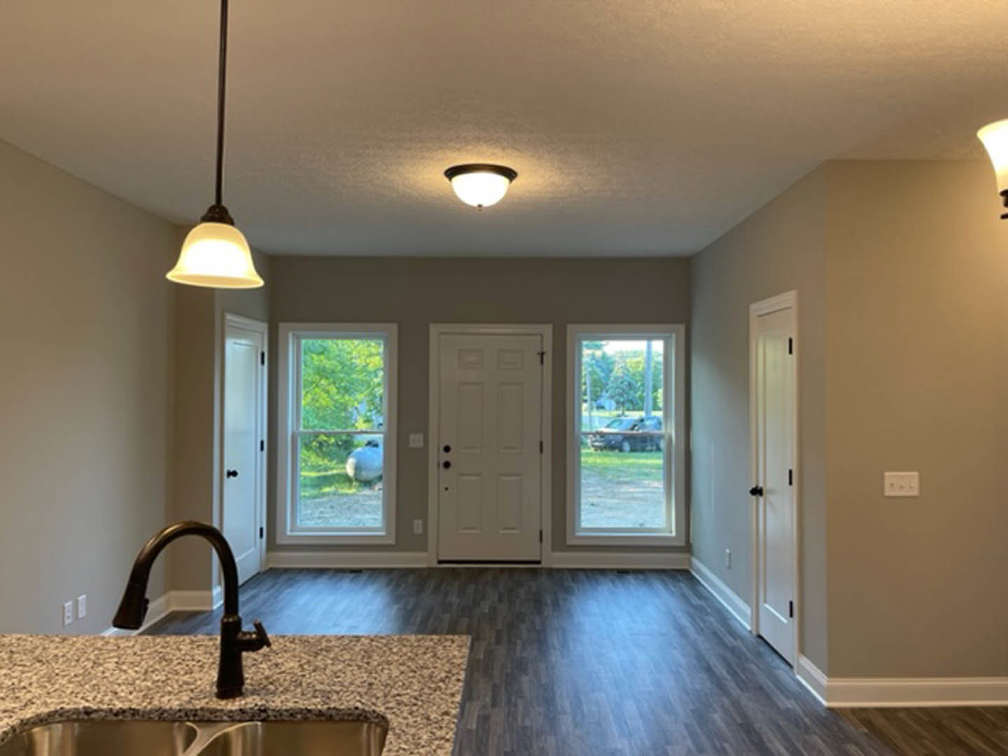 Open kitchen with wood flooring, white cabinetry, black faucet on a countertop, stainless steel sink, white door with black knobs, three-switch wall plate, large window showing