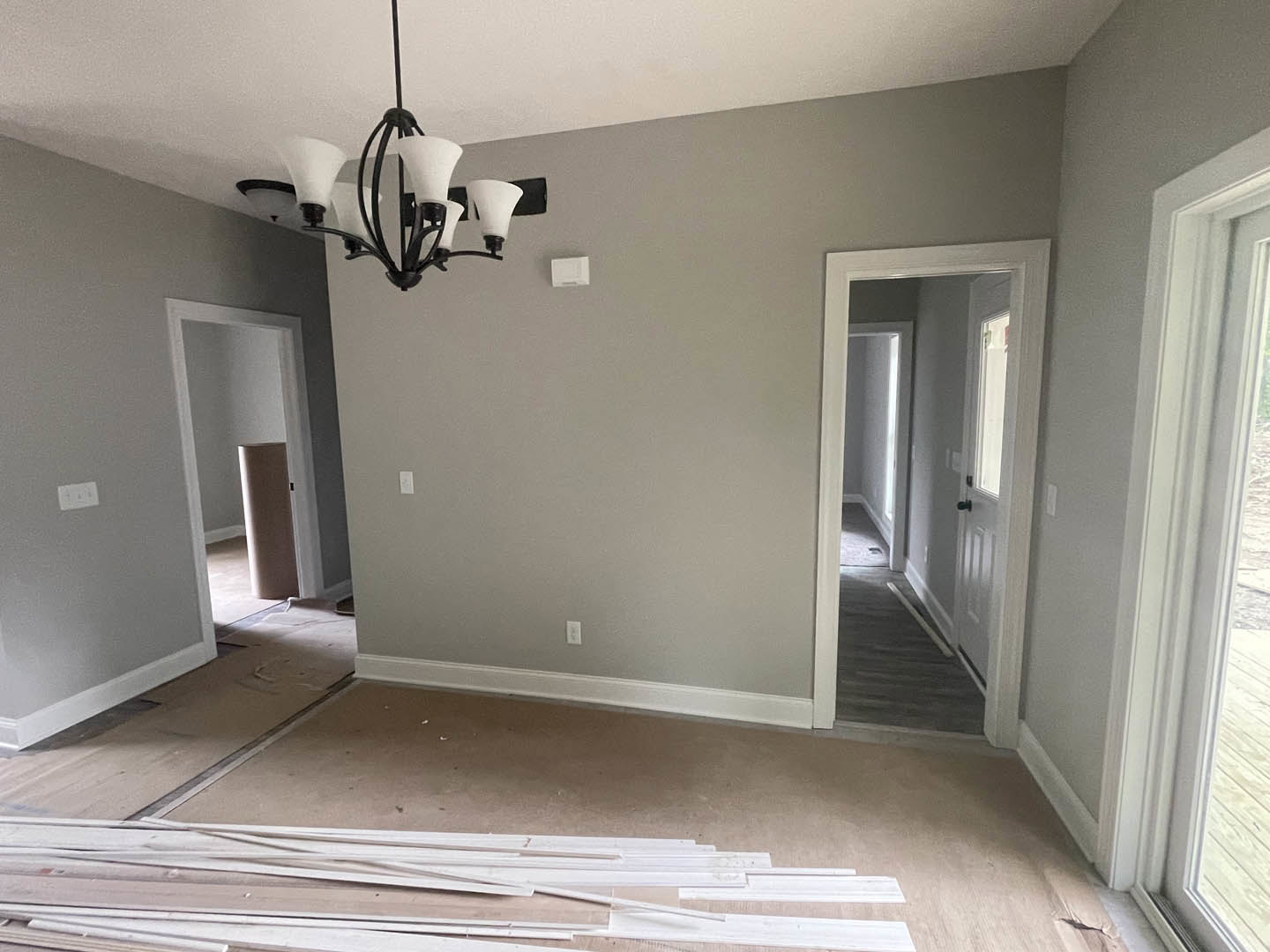 Carpeted living room with ornate chandelier, white pillar, and hallway leading to door and window; white wood planks stacked in foreground.