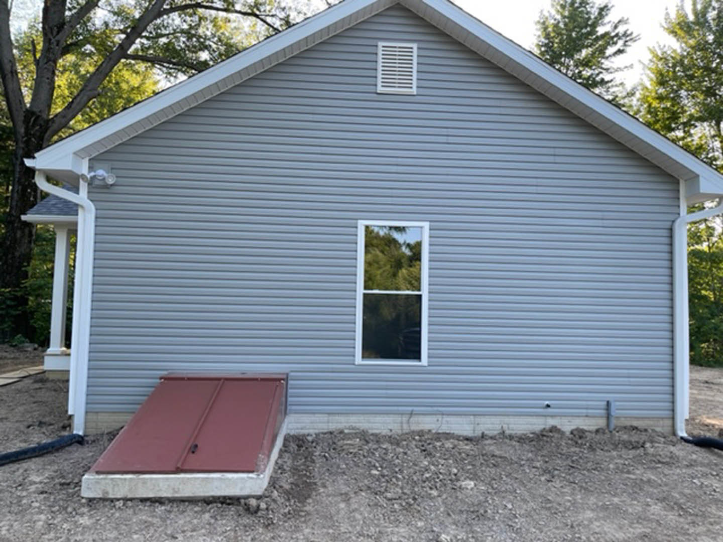 White cottage-style home with open front door, metal roof, ramp leading to entrance, large window reflecting trees, visible exterior vent, and landscaping with a pile of dirt in