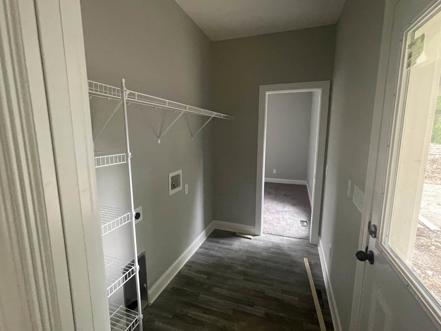 Hallway with dark wood flooring, white trim, built-in white shelf, open white door leading to a room with grey carpet, plaster walls, and ceiling