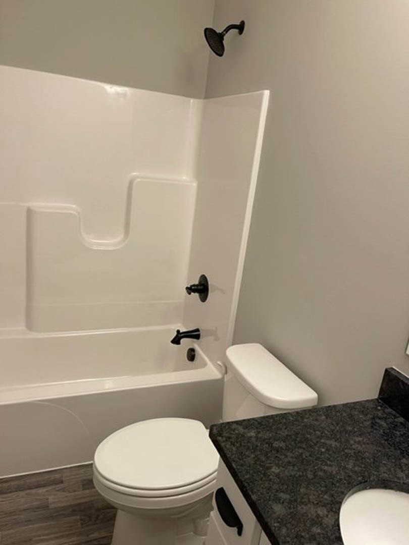 Modern bathroom featuring a white toilet beside a rectangular bathtub, black countertop with integrated white sink, light tile flooring, and neutral wall tiles.