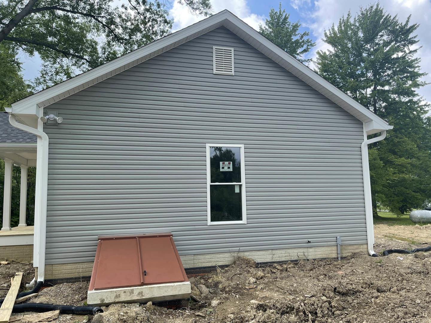 Grey siding house with brown metal door, single window displaying a sign, white vent above, concrete entry, grassy yard with large round object, trees and sky in background