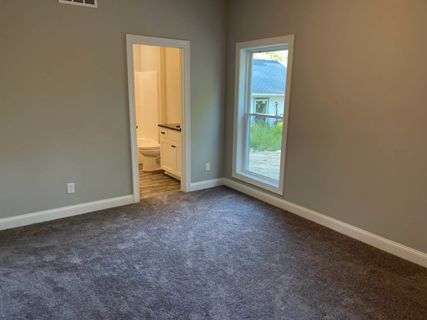 Open white door leading to carpeted room with large window, white trim, and neutral walls; partial view of bathroom with sink and bathtub visible.