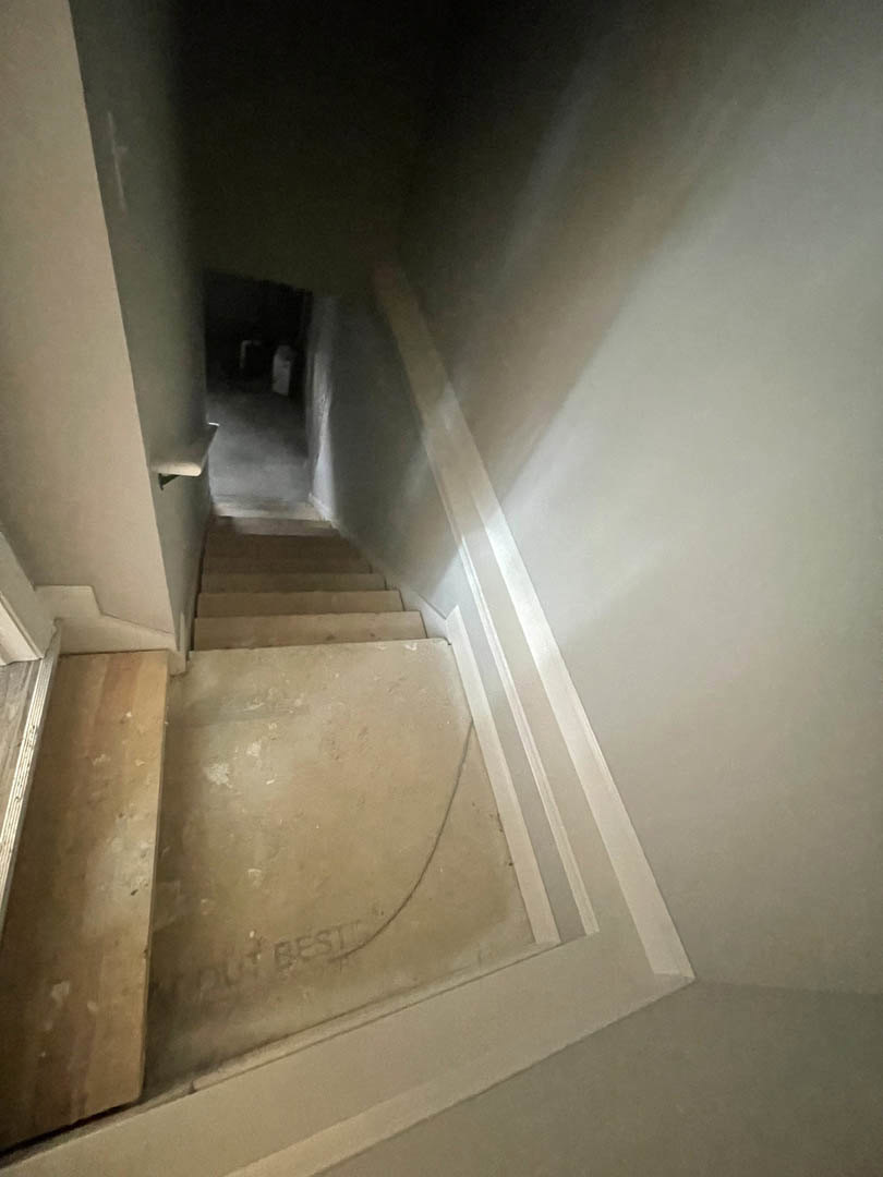 Wood plank staircase descending along a plaster-walled hallway, darkened with minimal lighting, chair visible at the end.