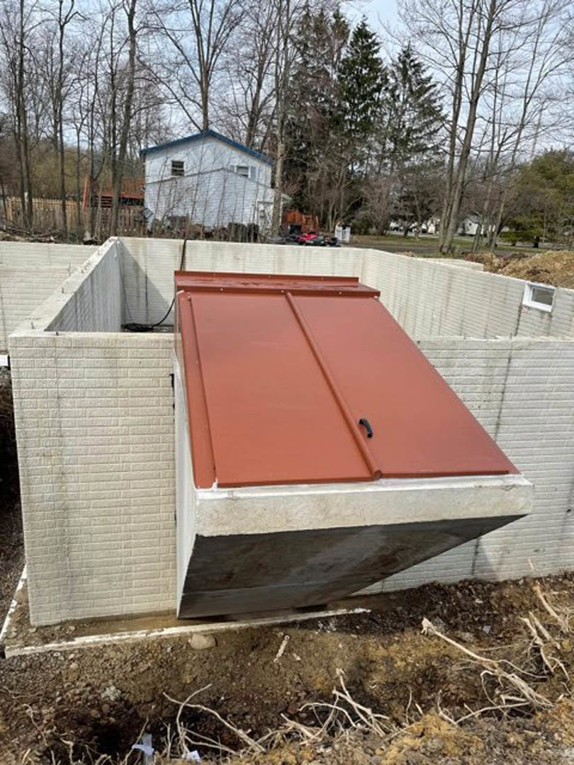 Red metal roof atop partially constructed home with exposed framing, white exterior walls, dirt ground, and surrounding trees.
