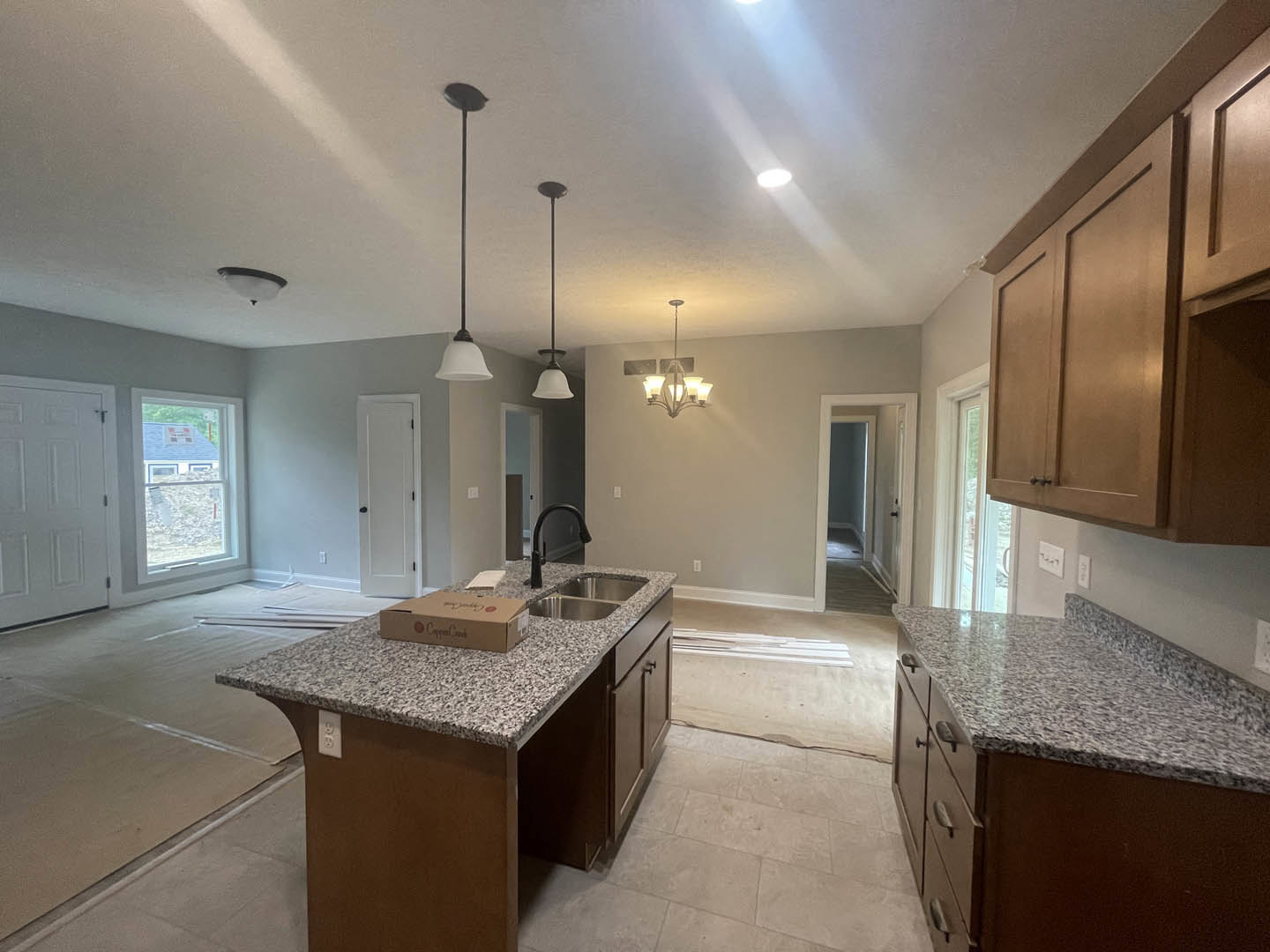 Granite countertops and tile backsplash in a kitchen with white cabinetry, stainless steel sink, kitchen island topped with a pizza box, and a window with blue roof visible outside